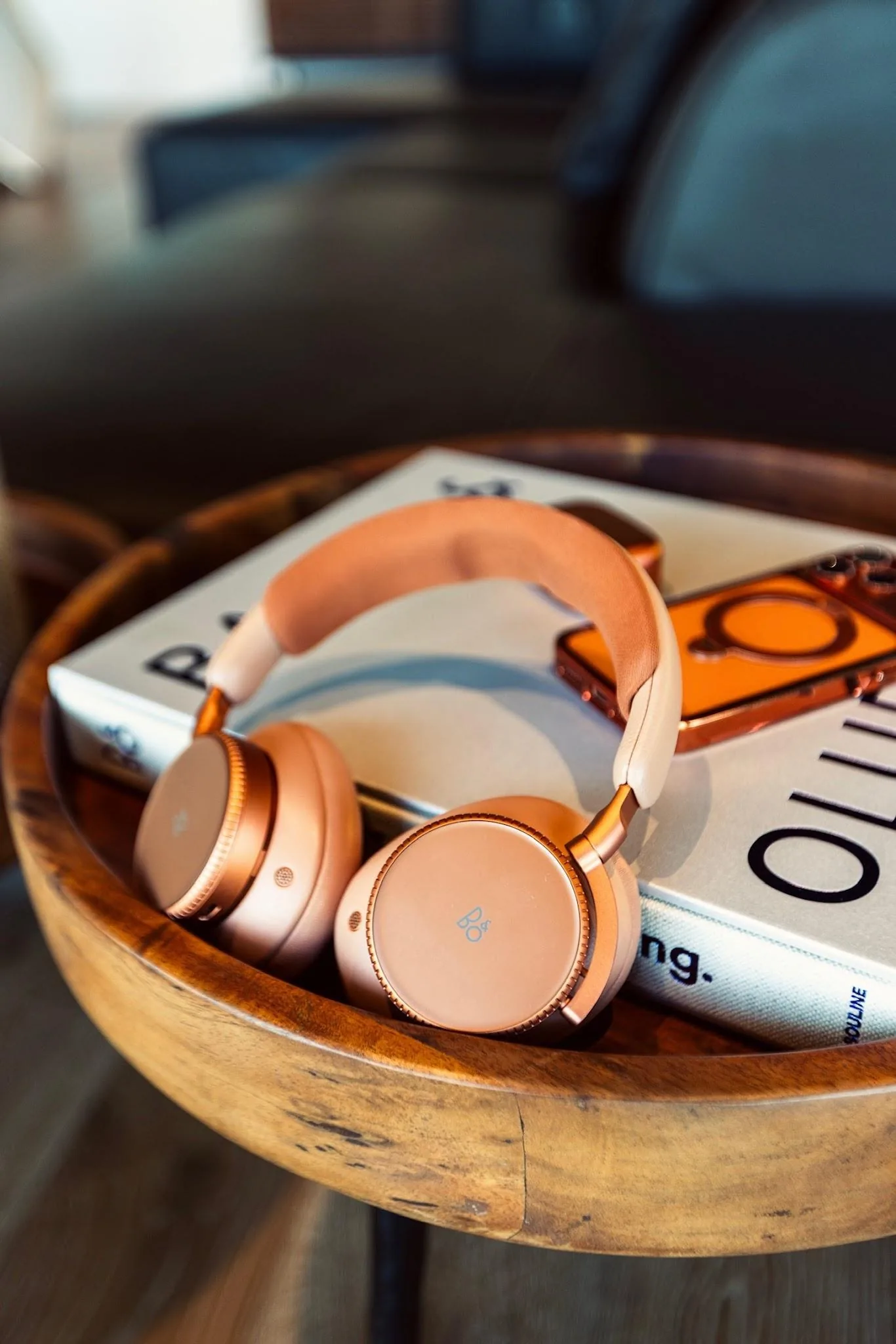 Copper-tone Bang & Olufsen Beoplay headphones resting on a Bang & Olufsen book on a round wooden side table