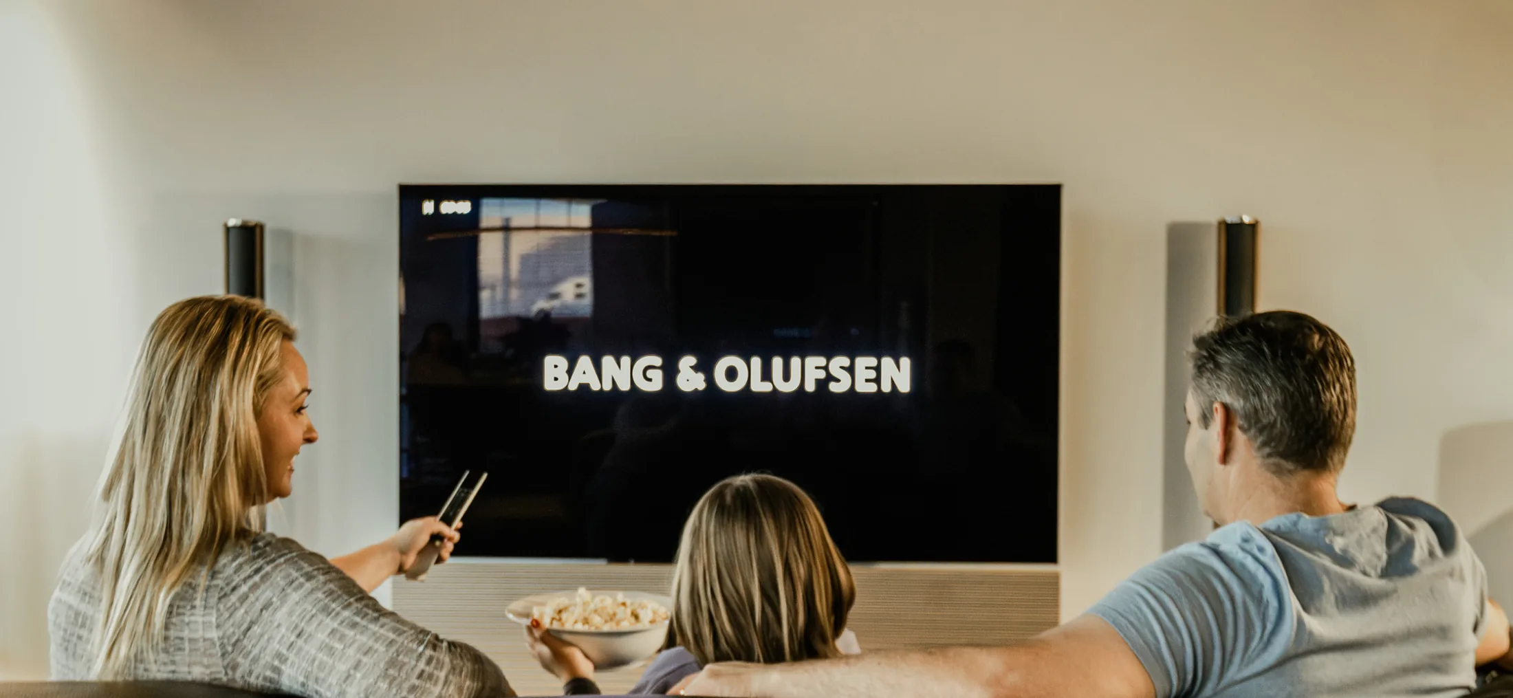 Family on a sofa watching a Bang & Olufsen television that shows the Bang & Olufsen wordmark, popcorn in hand