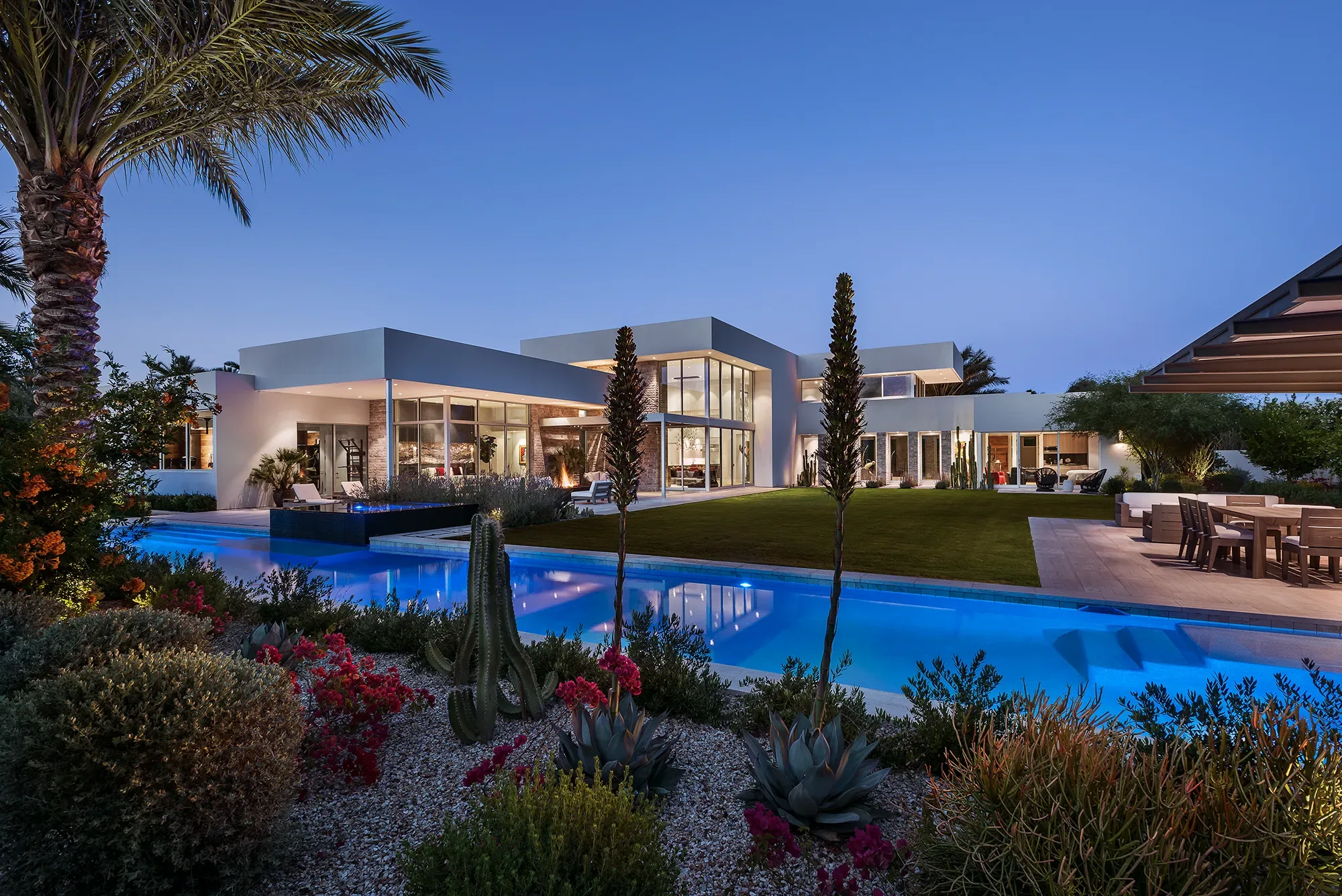 Rear exterior of a modern desert home at dusk, the pool and landscape lighting switched on