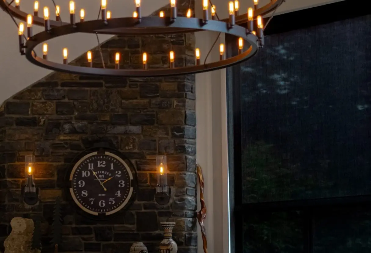 Great room of a client’s second home: a two-tier candle chandelier over a stone fireplace, with a motorized roller shade lowered across the tall window beside it