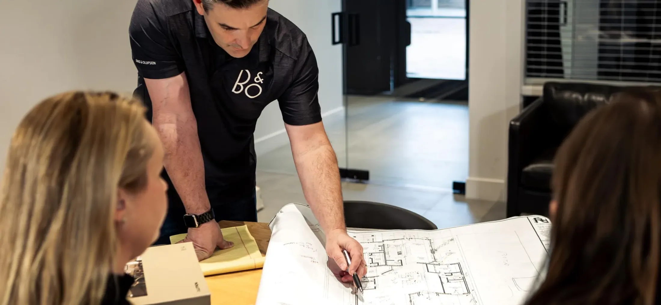 A Watt Integration team member in a Bang & Olufsen shirt pointing out details on floor plans to two clients