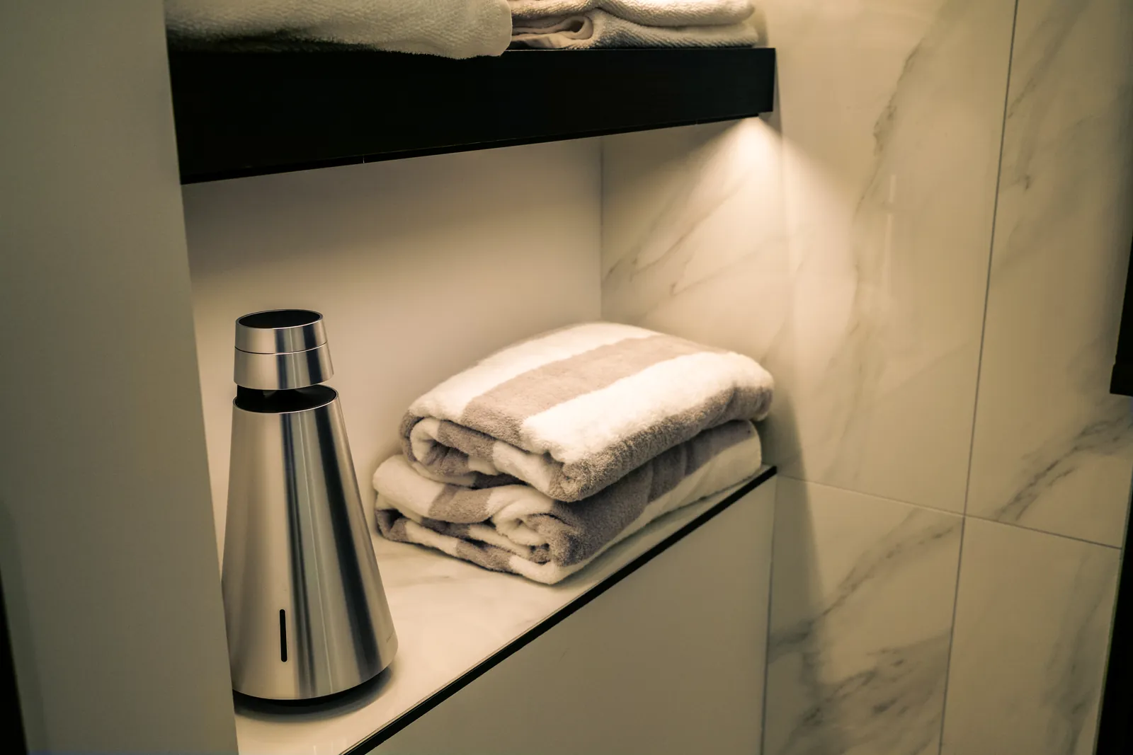 Bang & Olufsen Beosound 1 speaker on a marble bathroom shelf beside folded towels