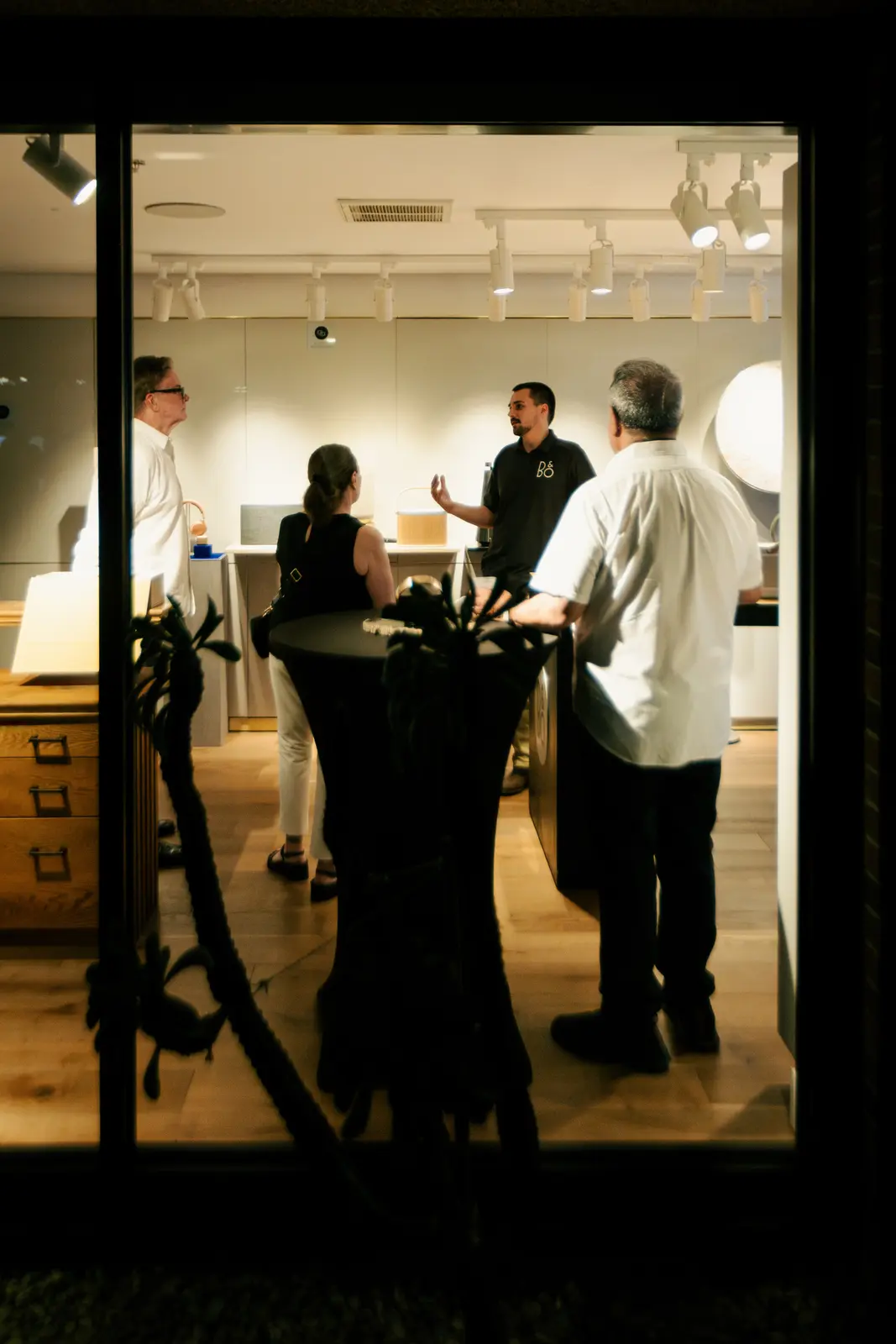Seen from outside at dusk, a Watt Integration team member talking with two visitors among the lit displays of the Scottsdale showroom