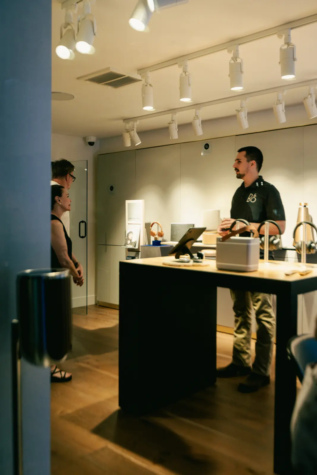 Inside the Bang & Olufsen Scottsdale showroom — a display island of speakers and headphones under track lighting, with visitors browsing