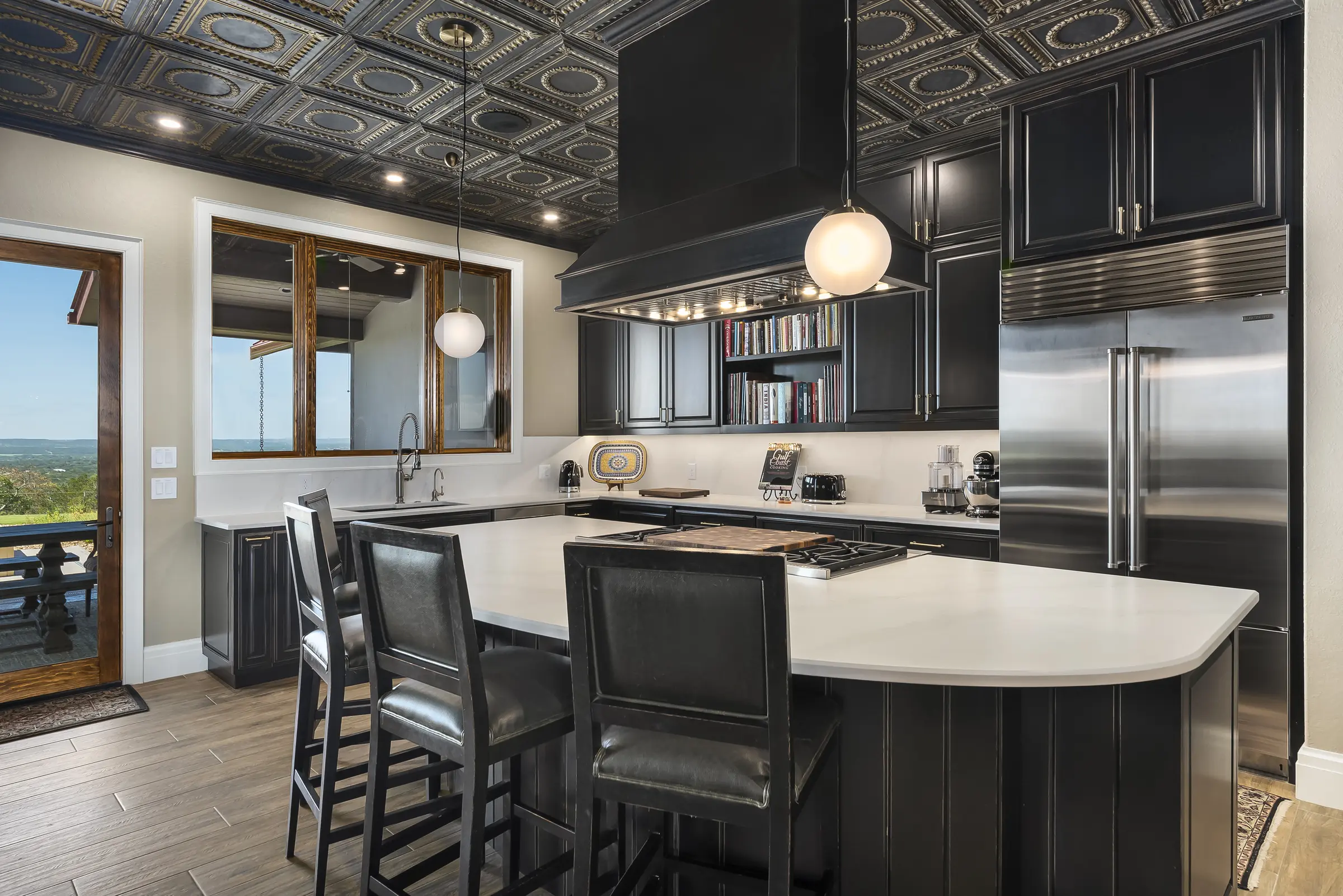 A kitchen with black cabinetry, a pressed-tin ceiling and a white stone island, opening to a window seat and a view beyond