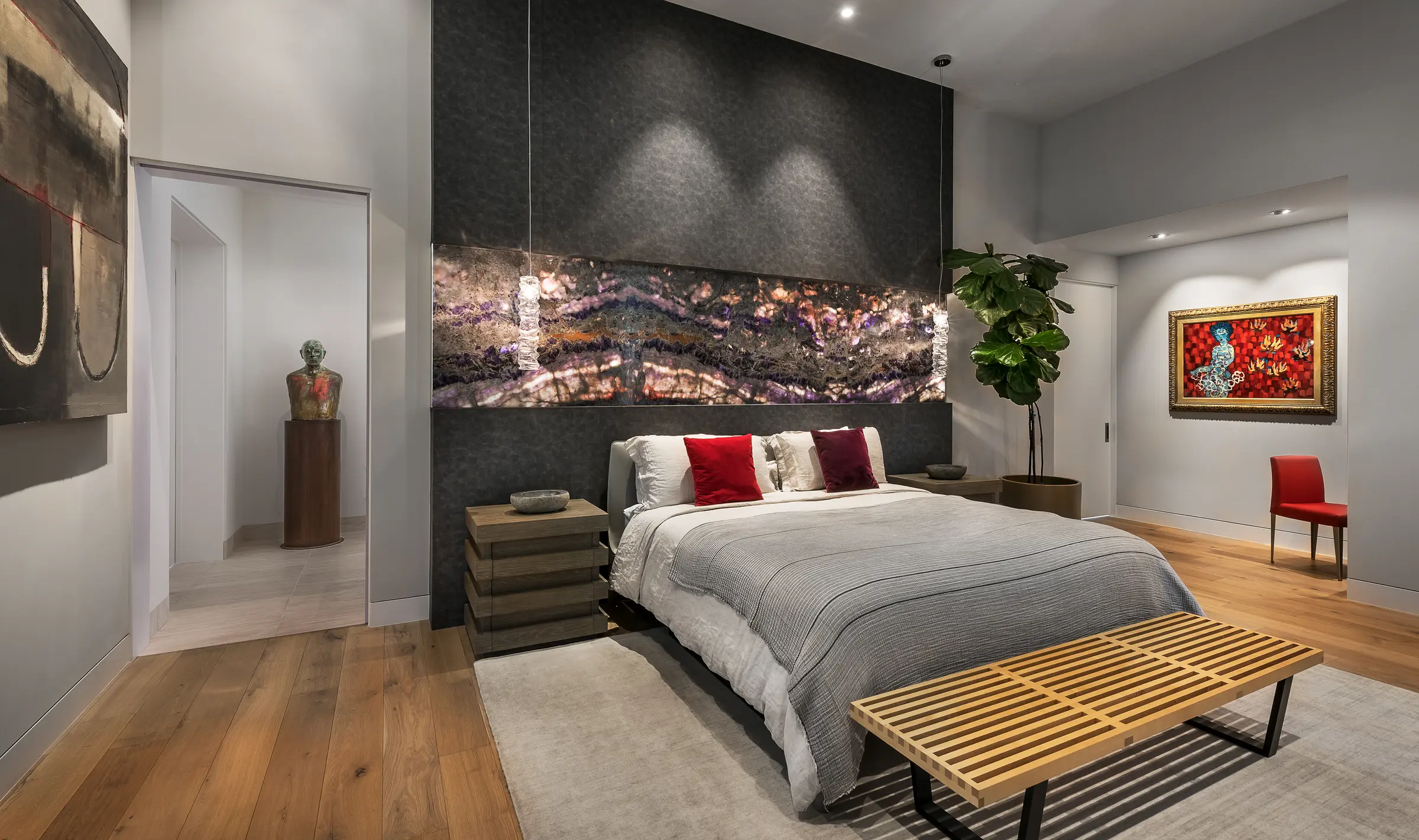 A primary bedroom with a backlit stone headboard panel, recessed downlights over the bed and framed art on the adjoining walls