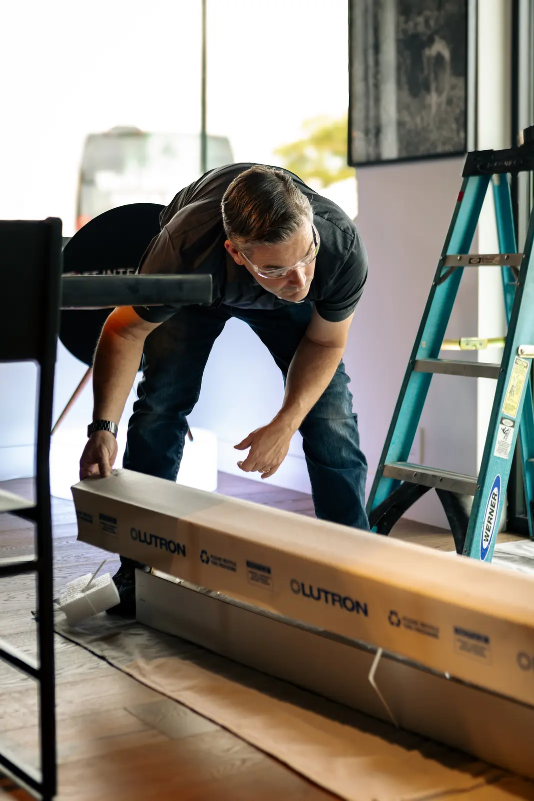 A Watt Integration installer lifting a long Lutron shade carton clear of its packaging on a protected floor, step ladder alongside