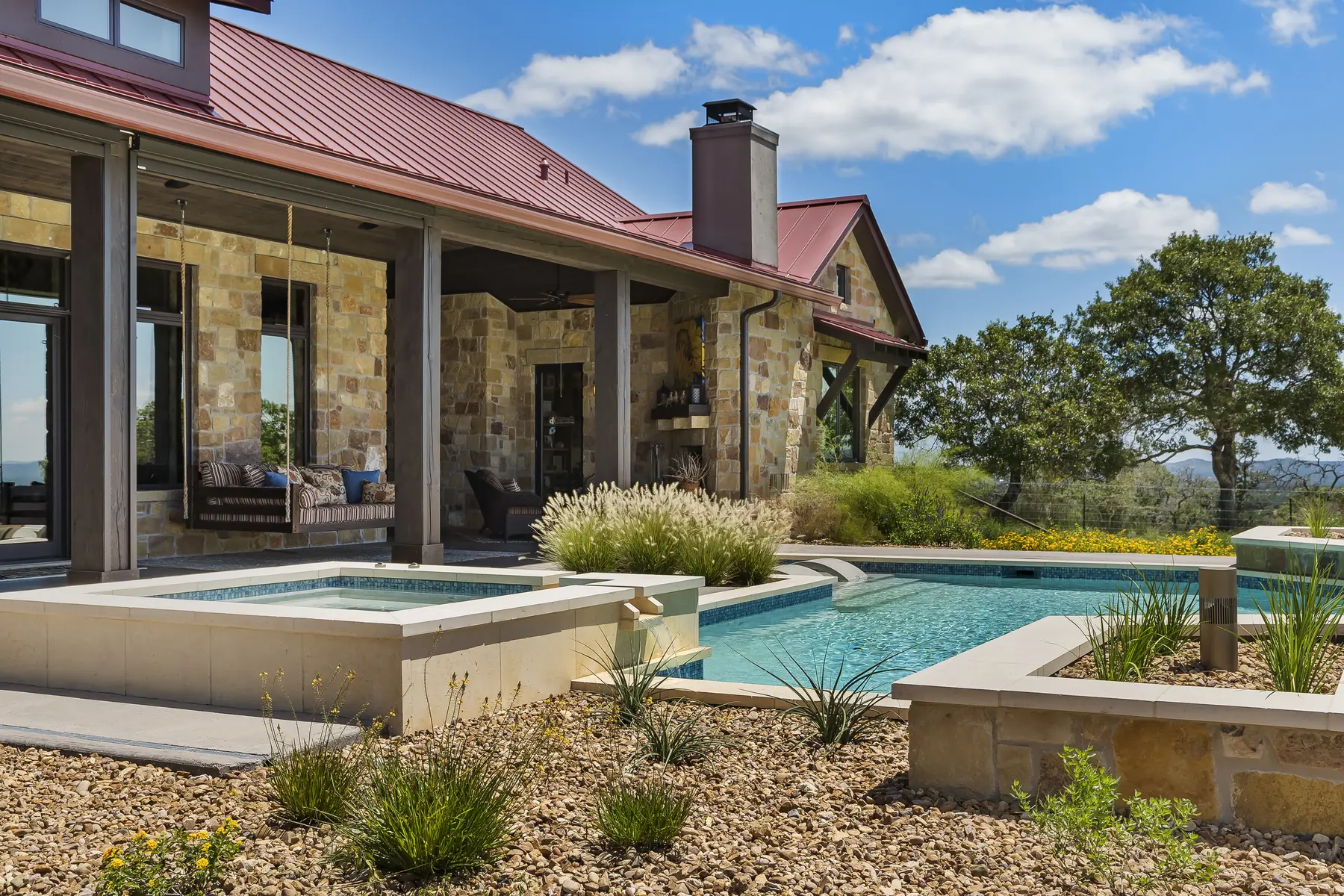 A stone home with a standing-seam roof beside its pool and raised spa, ornamental grasses along the terrace edge