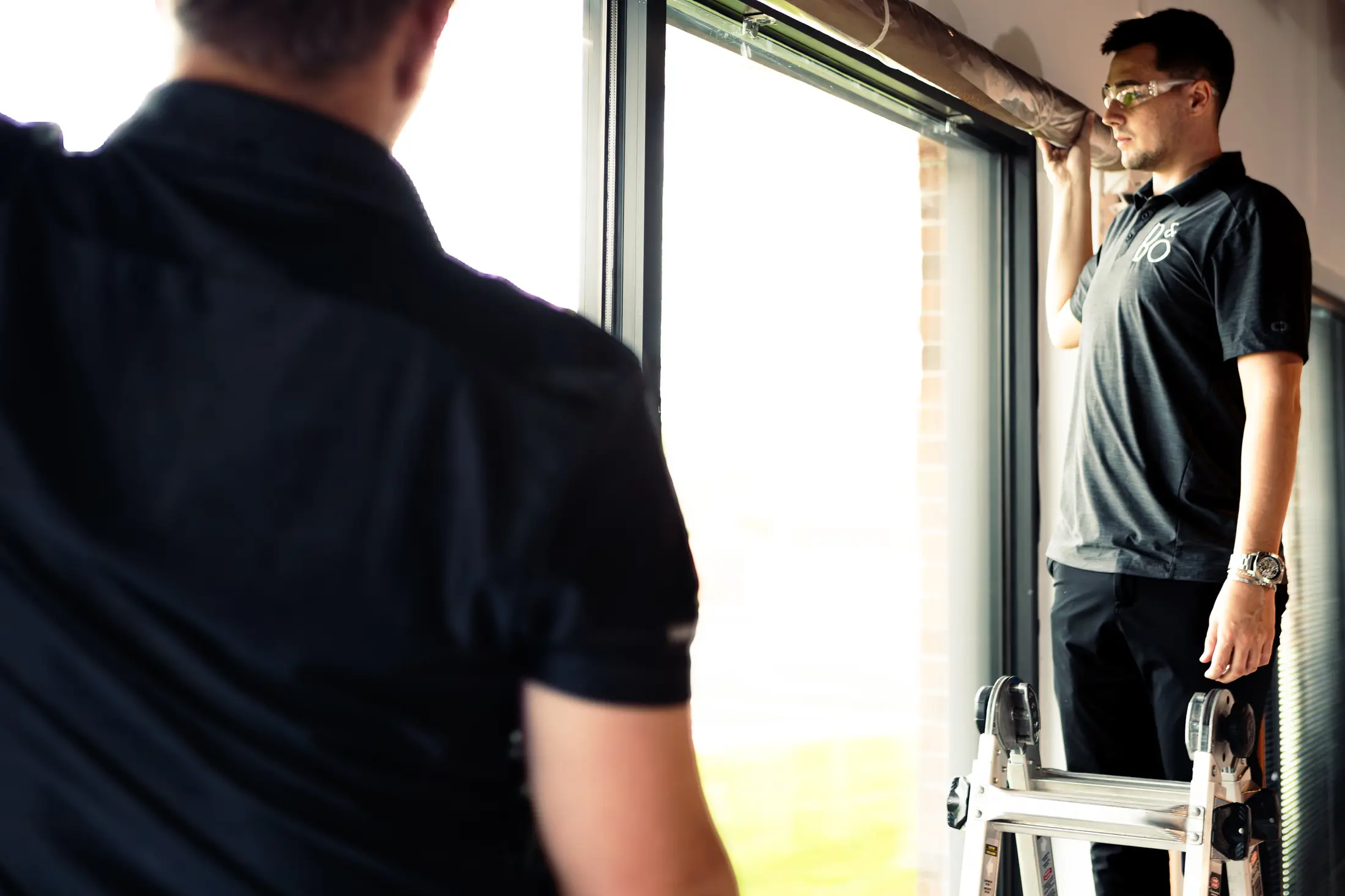Two Watt Integration installers fitting a shade tube into the head pocket above a tall window, one working from a step ladder
