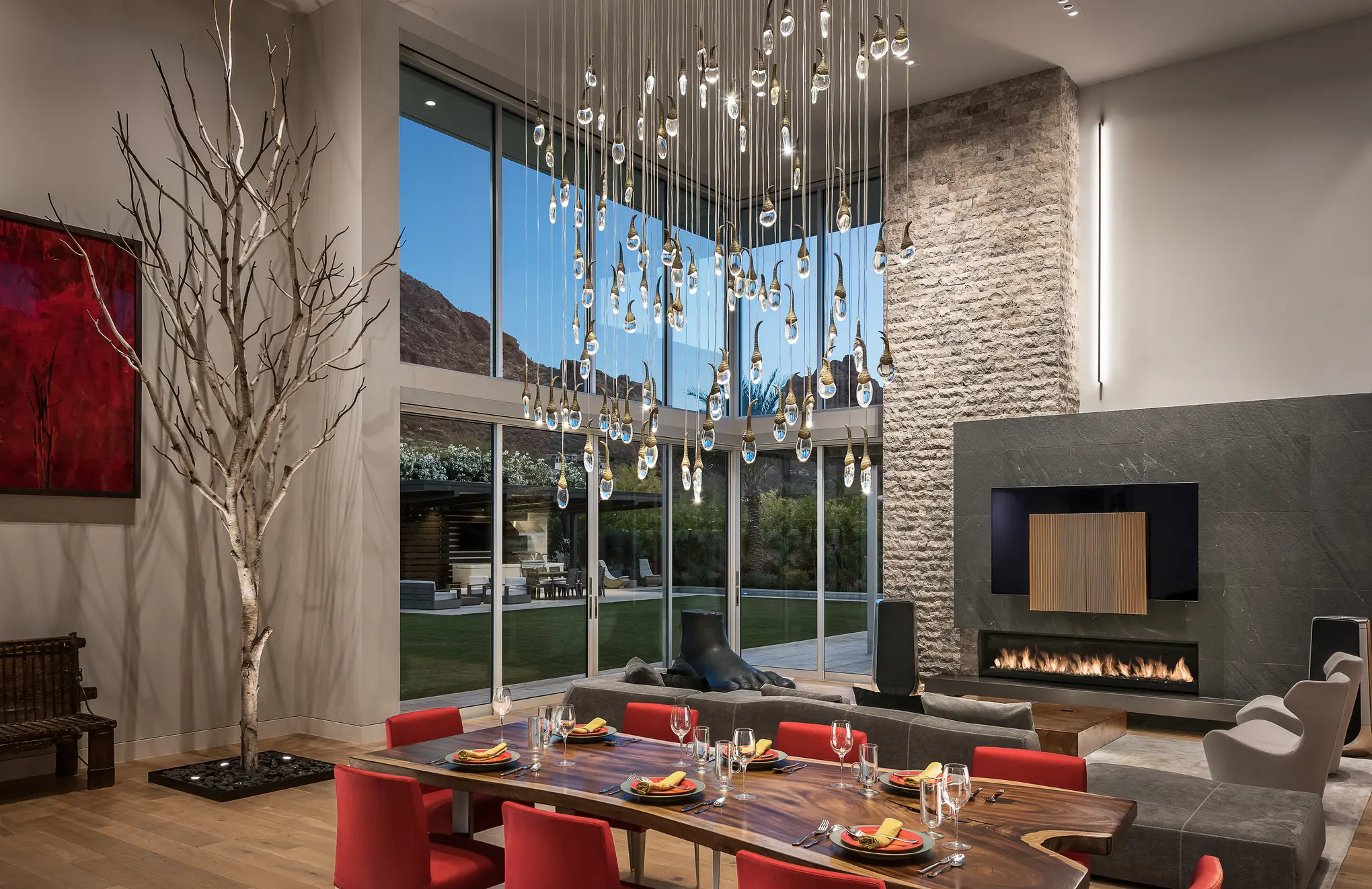 Dining and living space at dusk beneath a cascading glass-drop chandelier, with a fireplace and wall-mounted screen beyond
