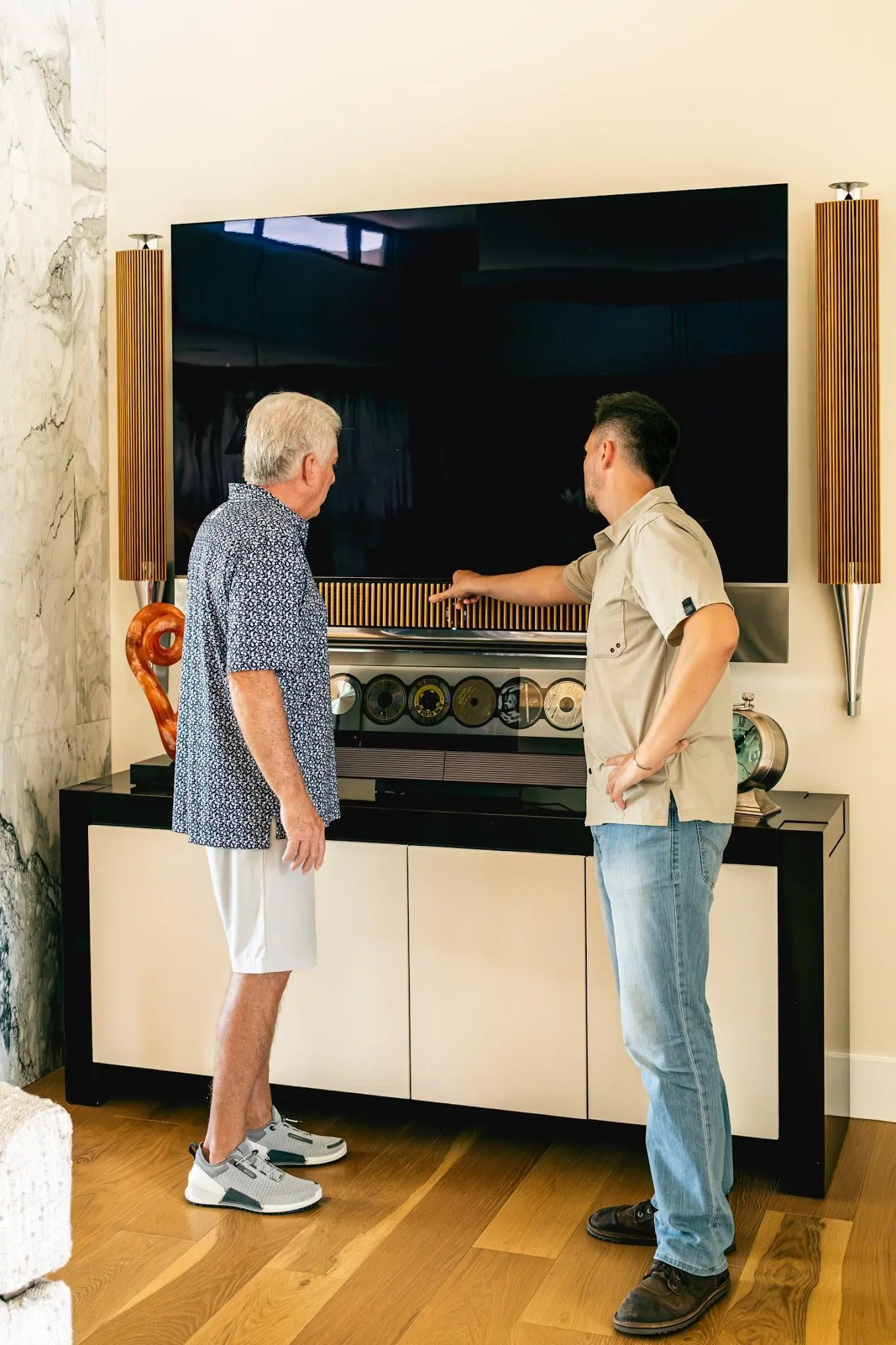 Two people at the media wall of a Scottsdale home — a wall-mounted television flanked by tower speakers above a lacquer console